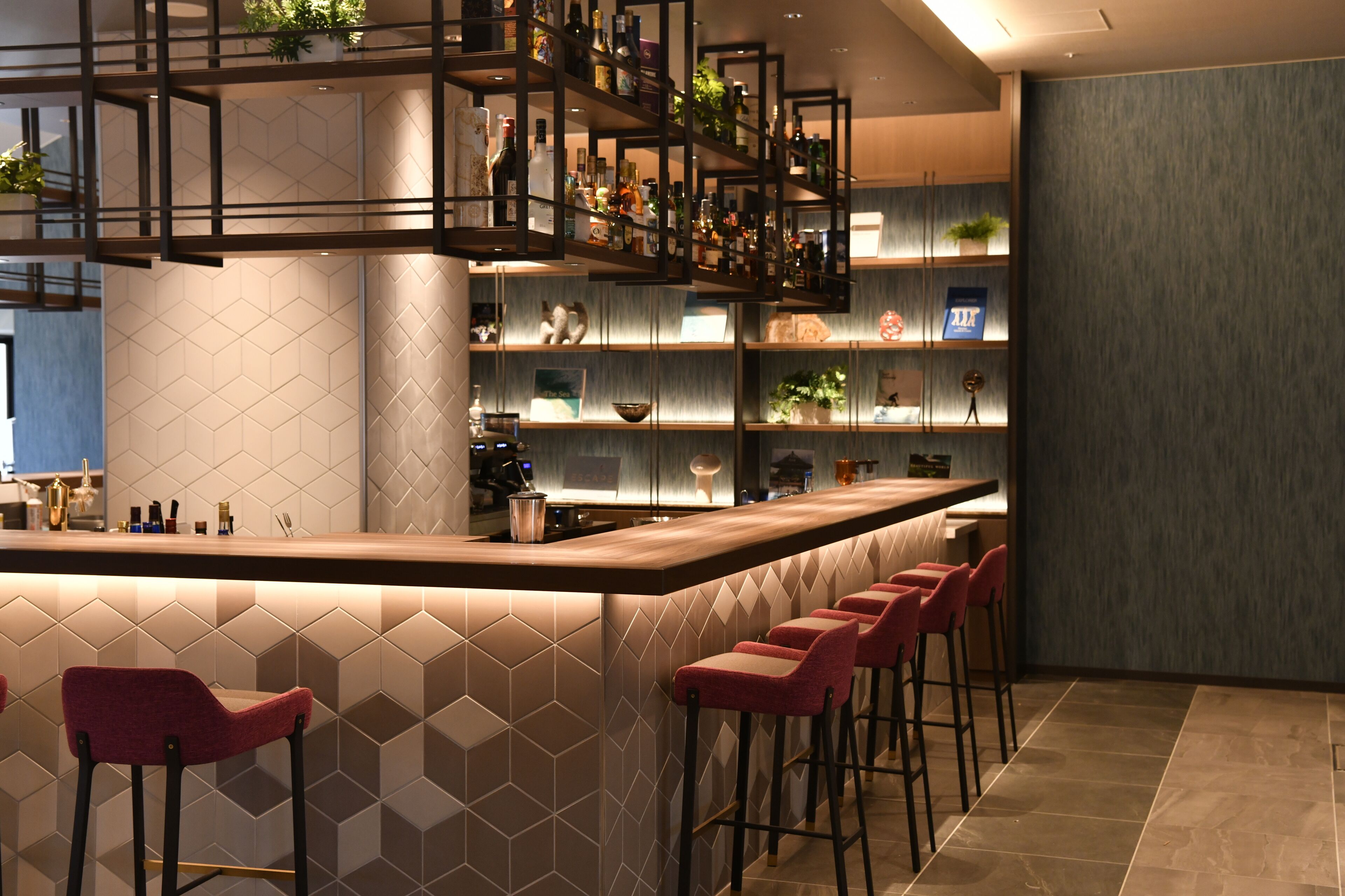 Un bar moderne doté d’un long comptoir, de plusieurs étagères remplies de bouteilles et de tabourets de bar rouges.