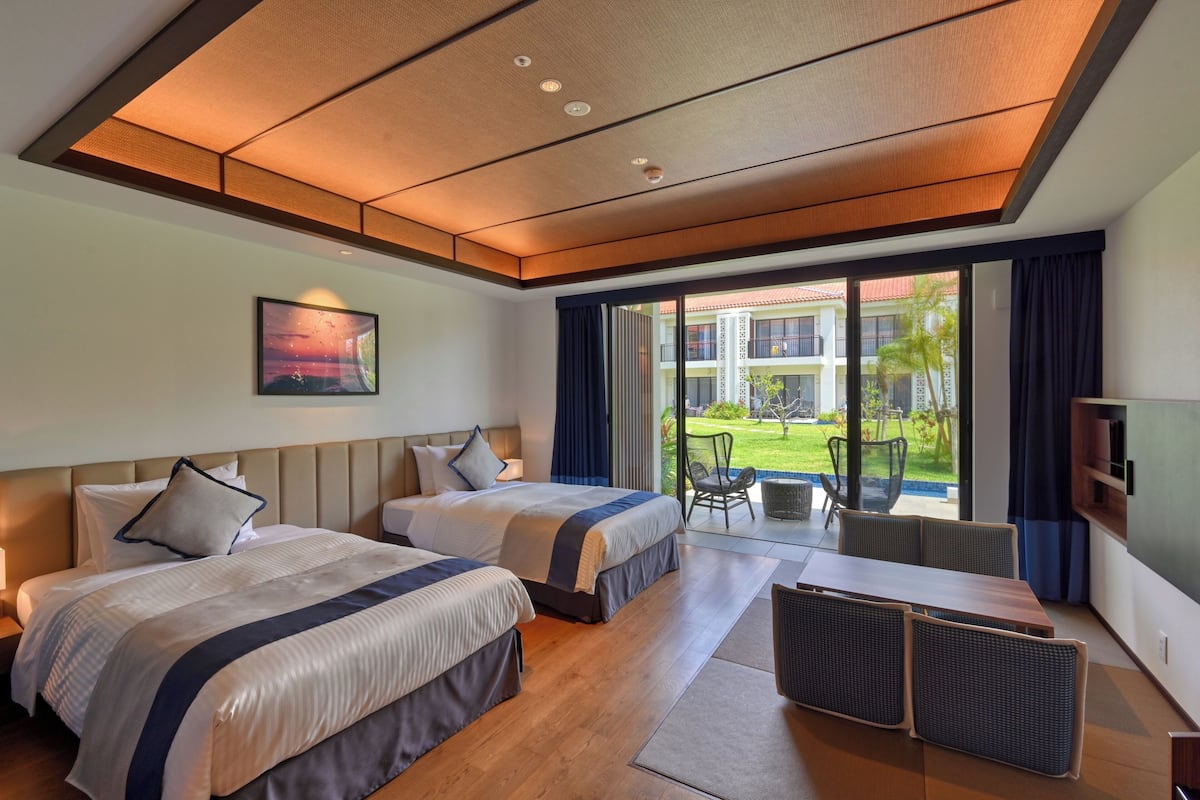 A hotel room with two beds, a dining table, and a view of the outdoor area.