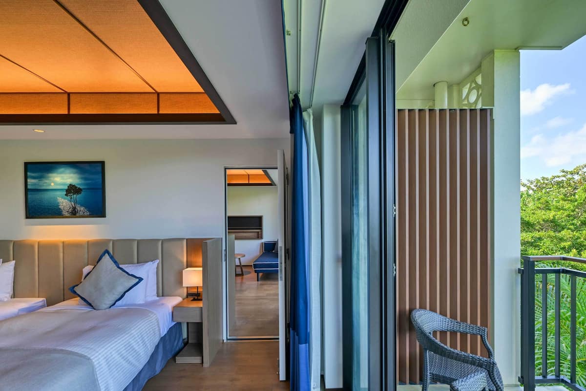 A hotel room with a bed, a desk, a chair, and a balcony with a view of greenery.