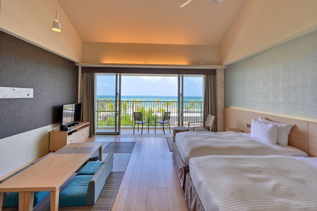 A hotel room with a bed, a desk, a TV, a balcony with a view of the ocean, and a ceiling fan.