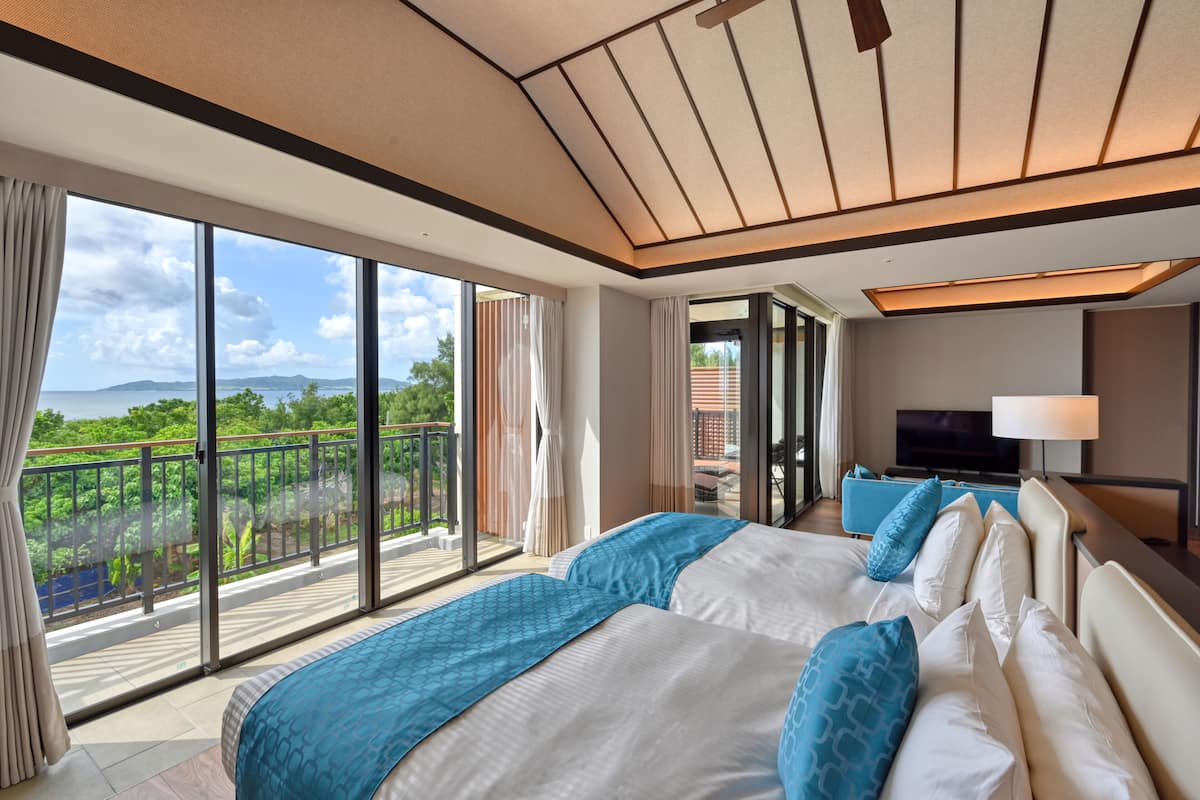 A spacious hotel room with two large beds, a balcony with a view of greenery and mountains, and a television on the wall.