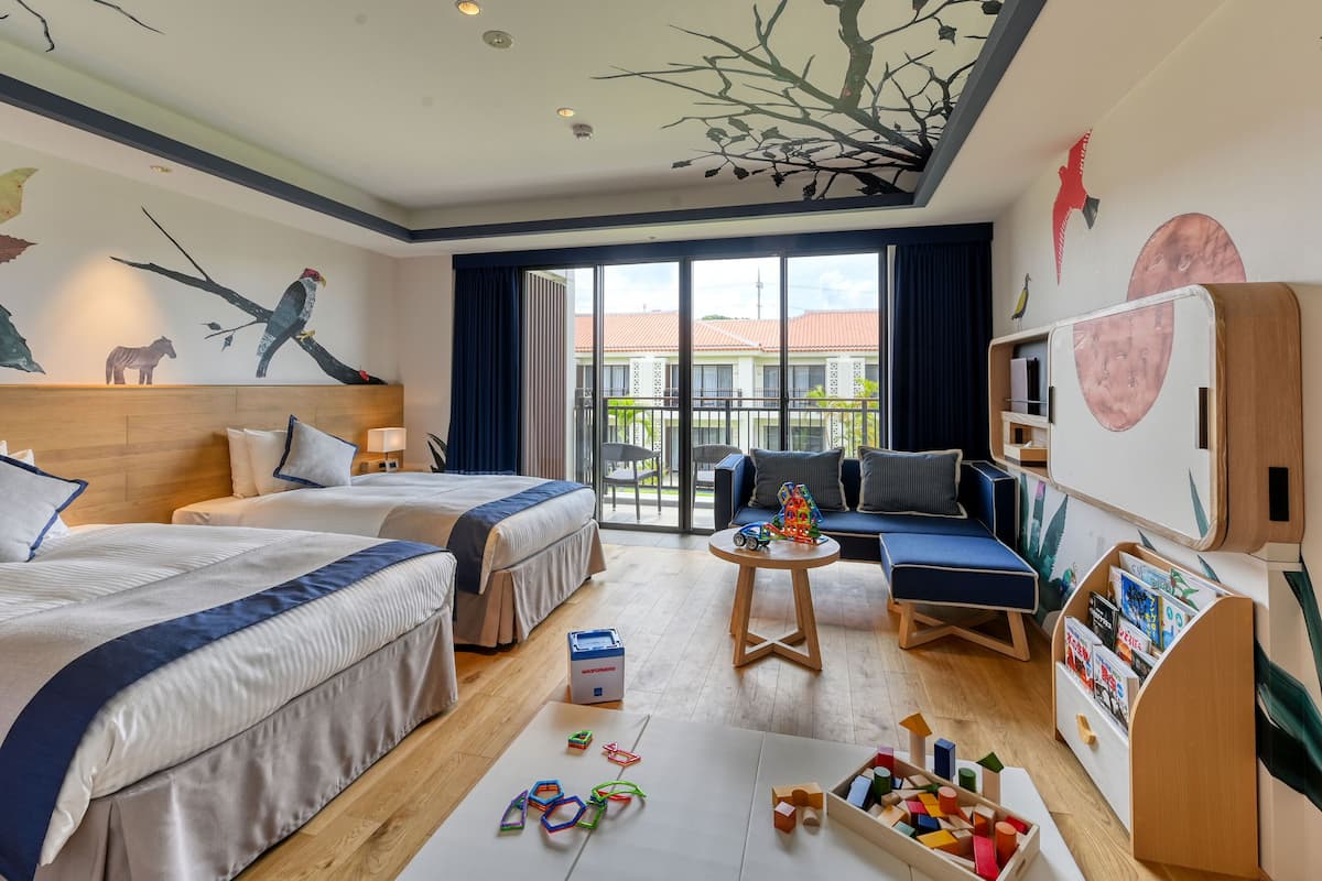 A hotel room with two beds, a sofa, a coffee table with toys, and a bookshelf.