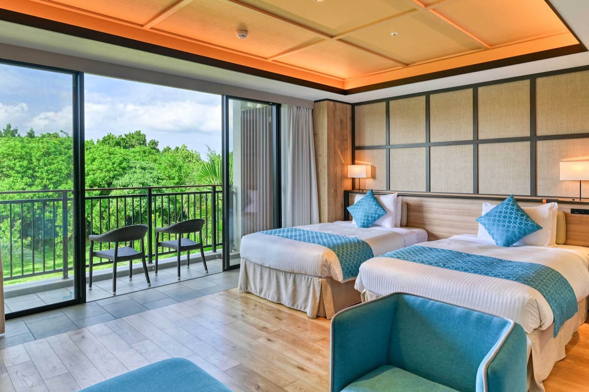 A hotel room with two beds, a balcony with chairs, and a view of greenery.