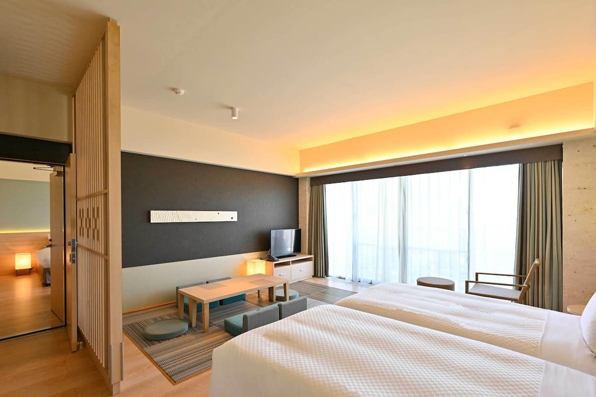 A hotel room with a large bed, a desk with a chair, a television, and a large window with curtains.