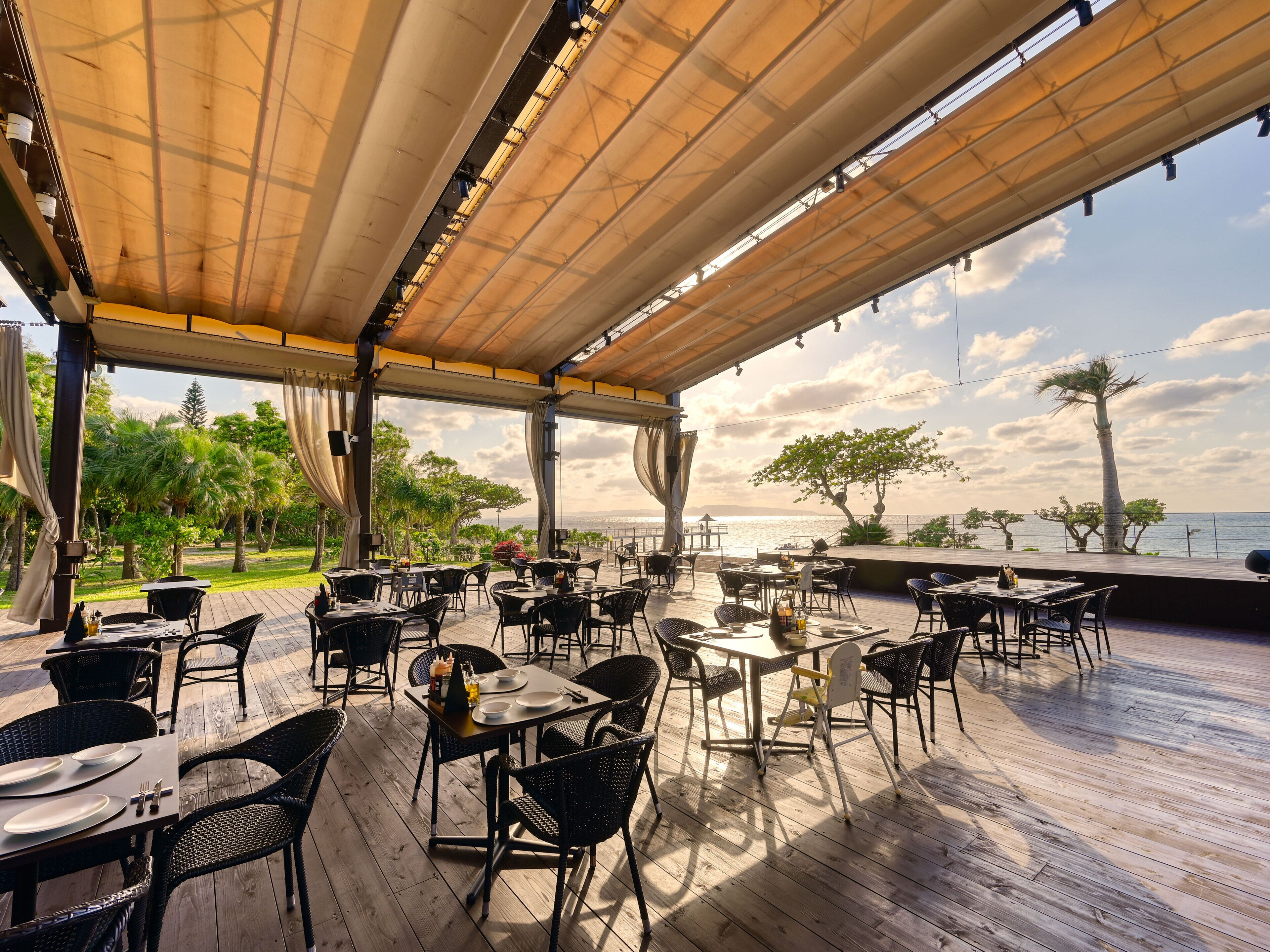 An outdoor dining area with tables and chairs, a wooden deck, and a view of the sea.