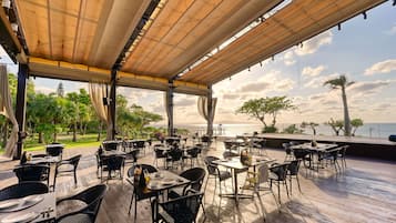 An outdoor dining area with tables and chairs, a wooden deck, and a view of the sea.