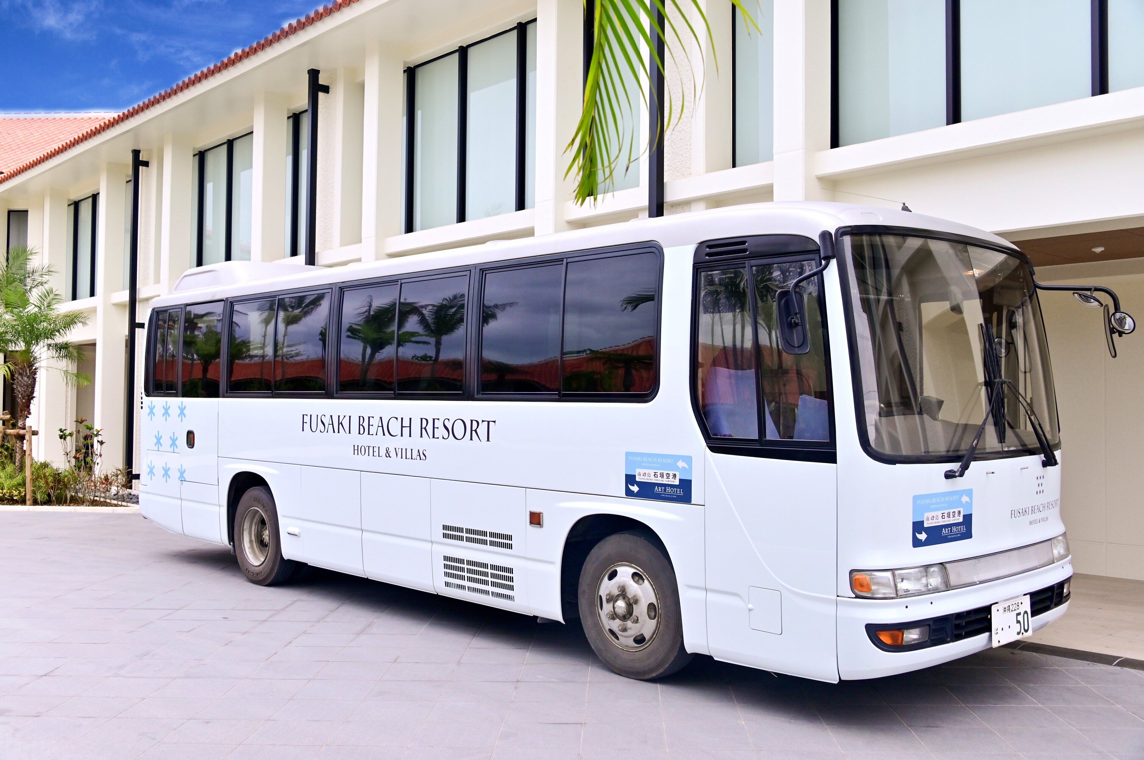 Un autobus bianco con la scritta "FUSAKI BEACH RESORT HOTEL & VILLAS" parcheggiato davanti a un edificio con tetto in tegole e ampie finestre.