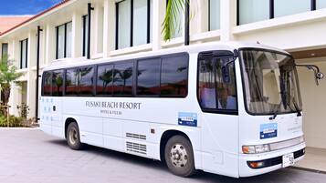 Un autobus bianco con la scritta "FUSAKI BEACH RESORT HOTEL & VILLAS" parcheggiato davanti a un edificio con tetto in tegole e ampie finestre.