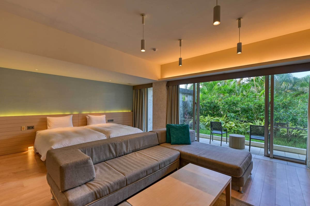 A modern hotel room with a large bed, a grey sofa, a small table, and a view of lush greenery through large windows.