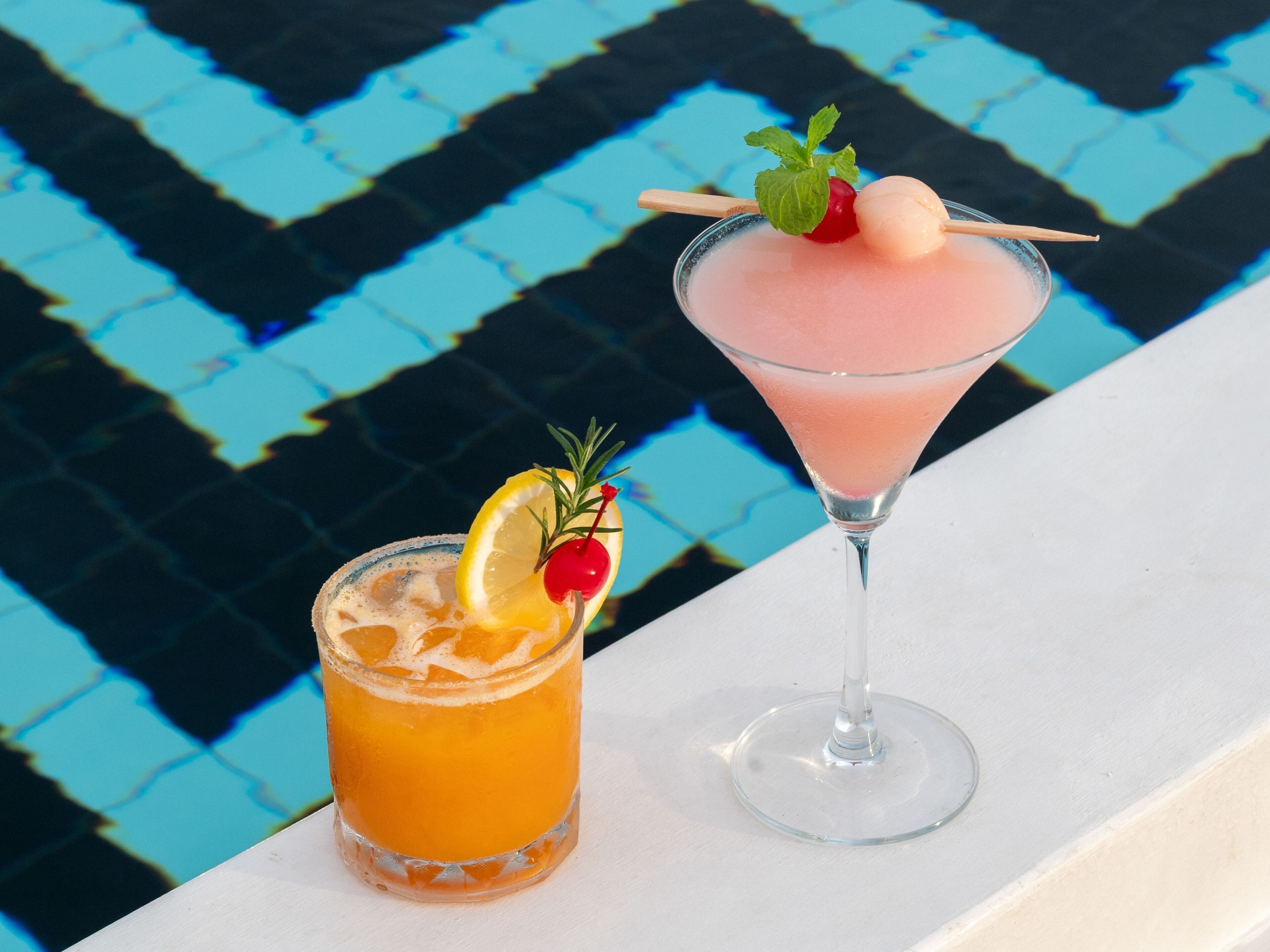 Two drinks, one in a short glass and one in a martini glass, with garnishes and a pool in the background.