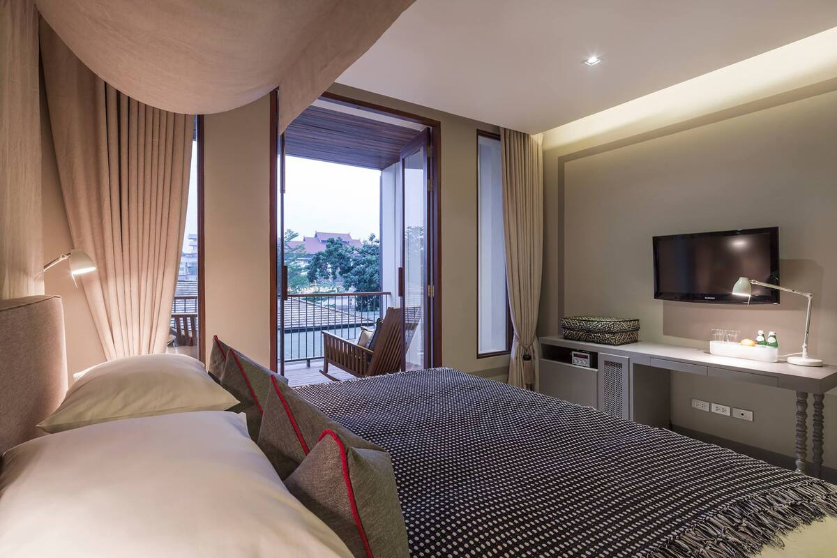 A hotel room with a large bed, a desk with a TV, and a balcony with a chair.