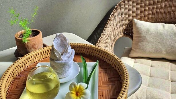 Ein Korbstuhl mit Kissen, ein Tablett mit einem Glas gelber Flüssigkeit, ein weißer Teller mit einer Blume und ein Blumentopf.