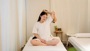 Eine Person sitzt auf einem Bett in einem Zimmer mit einem kleinen Tisch und einem Topfpflanzen.