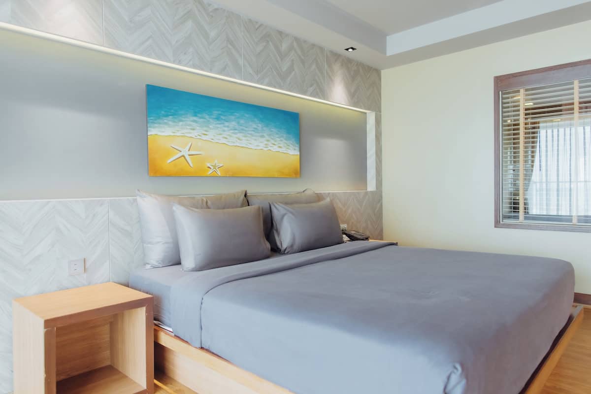 A modern bedroom with a bed, nightstands, a painting of a beach scene, and a window with blinds.