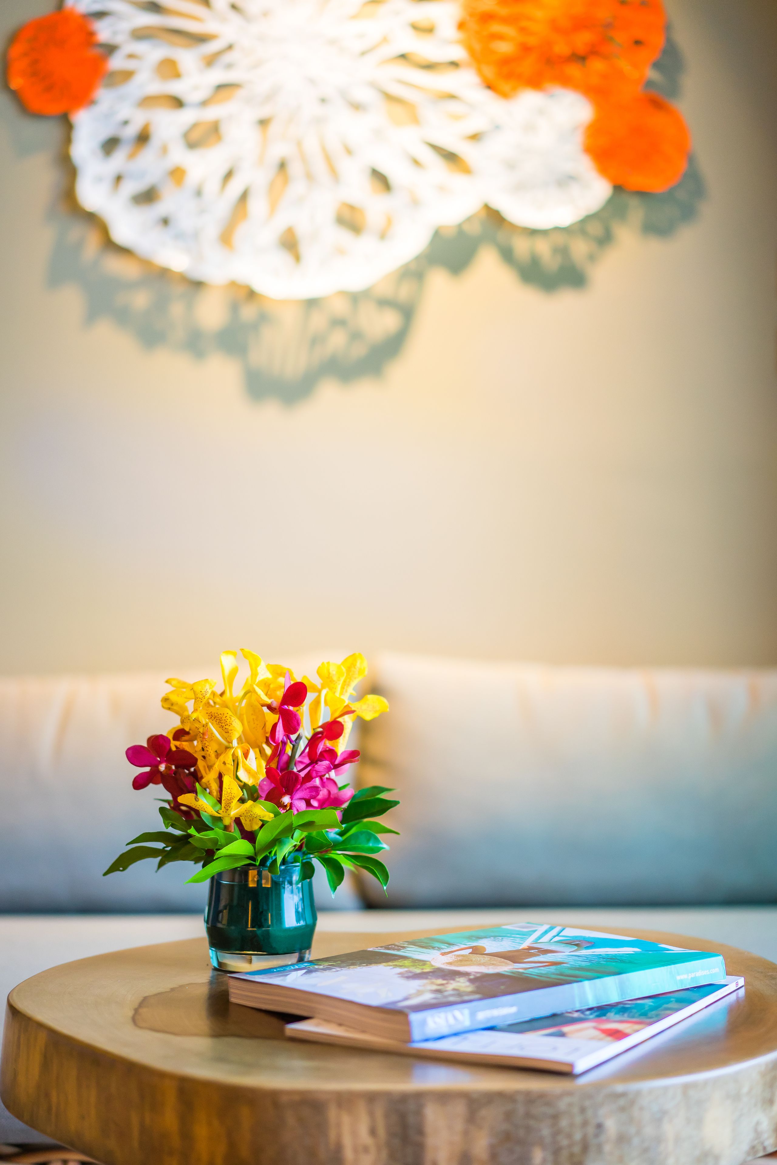 Une table ronde en bois sur laquelle se trouvent un petit vase en verre contenant des fleurs aux couleurs vives, une pile de magazines et un livre.