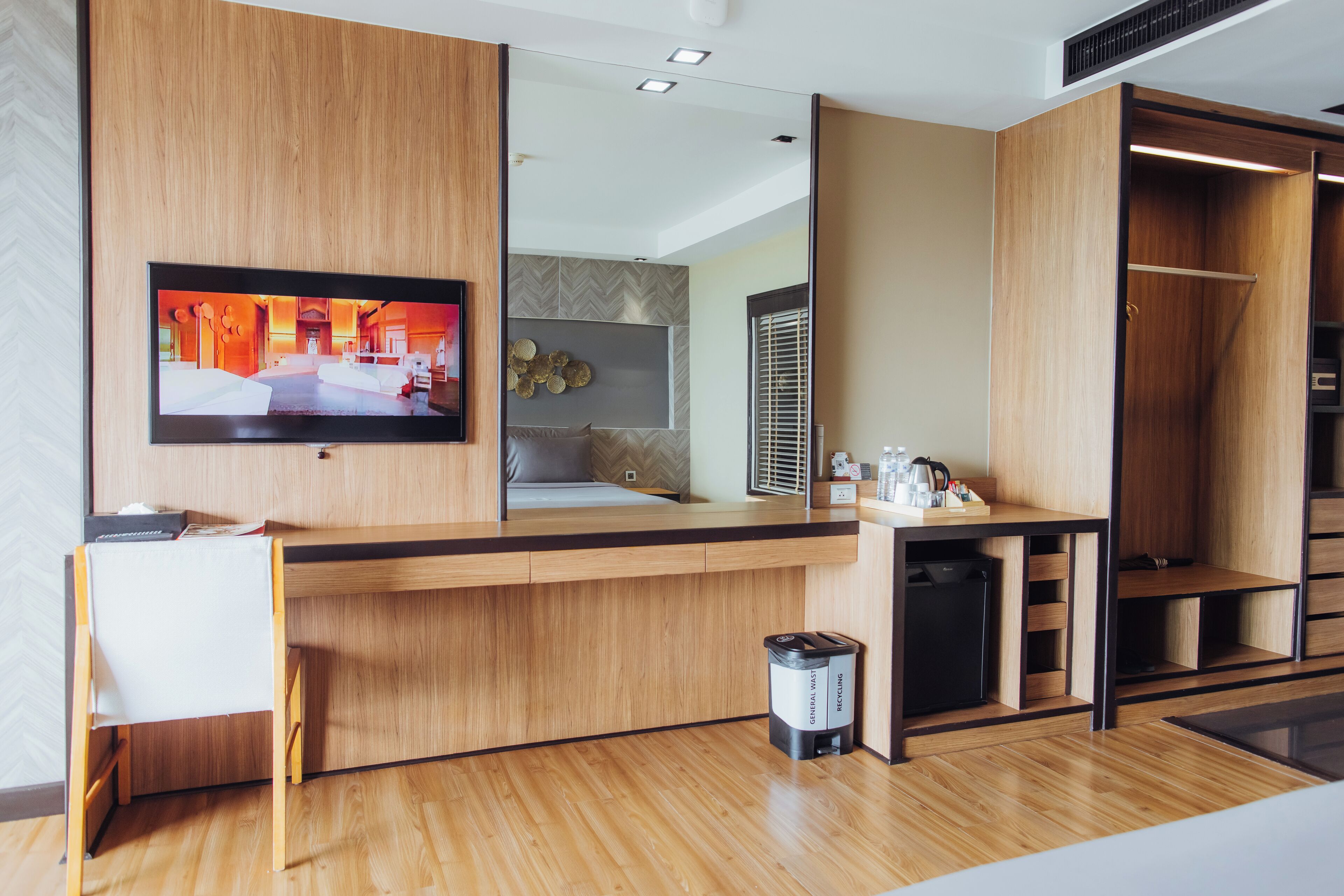 A modern hotel room with a wooden reception desk, a television mounted on the wall, a small trash bin, and a compact refrigerator.
