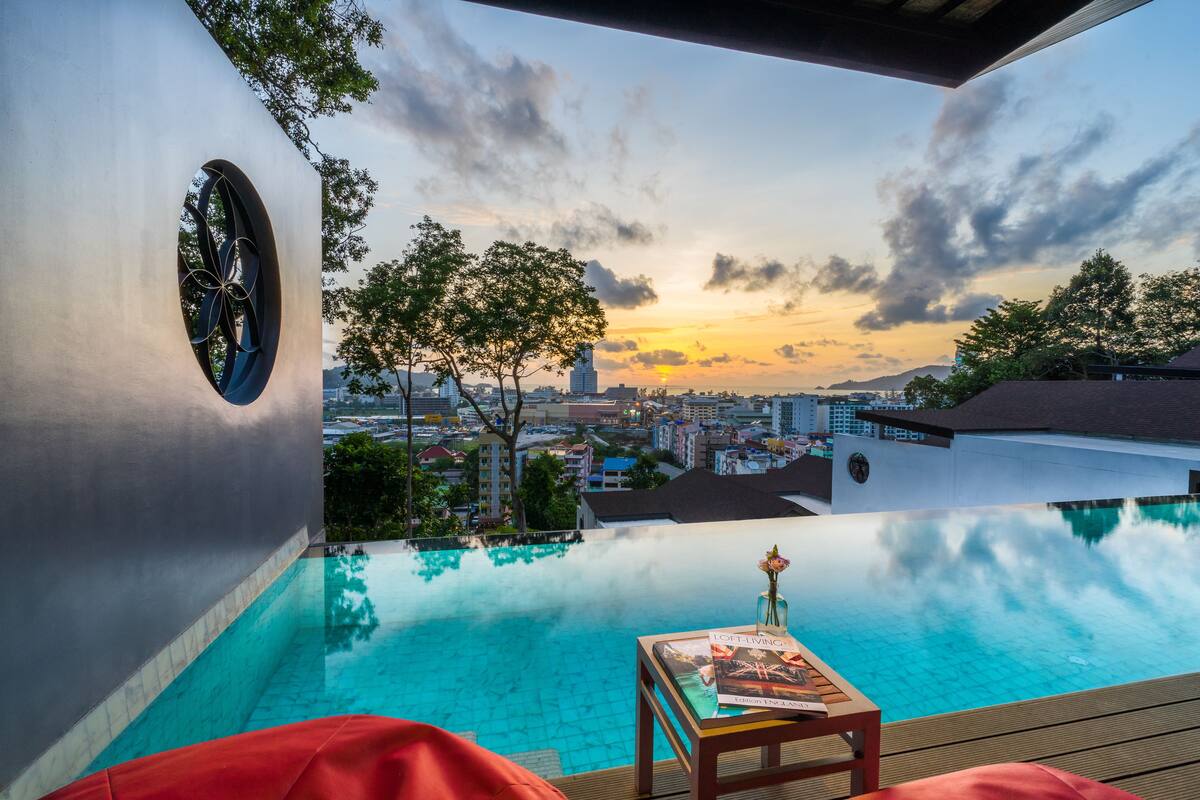 An infinity pool with a city view, a table with a magazine, and a red chair.