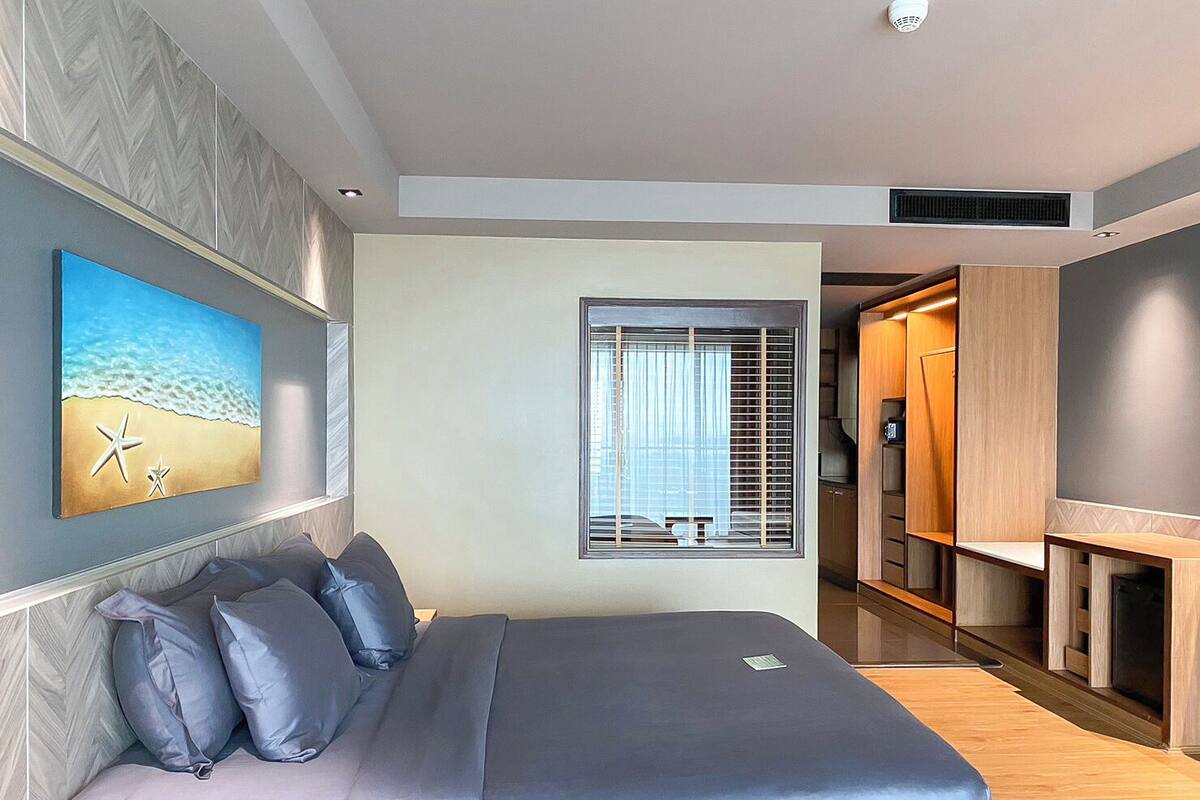 A modern bedroom with a bed, a beach-themed painting, a window with blinds, and a walk-in closet.