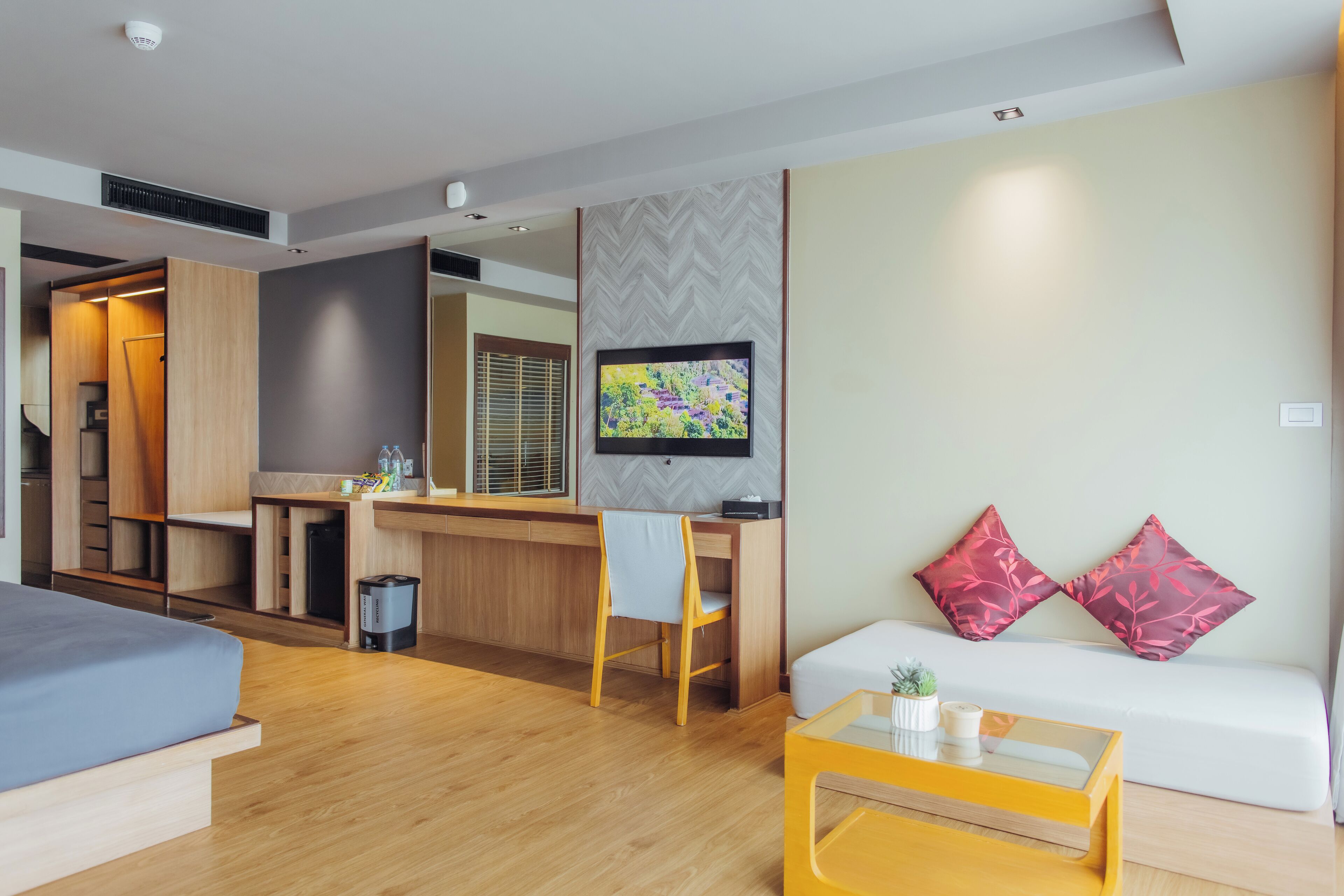 Une chambre d’hôtel moderne avec un plancher en bois, un canapé avec des coussins rouges, une petite table avec une plante, un téléviseur fixé au mur et un coin cuisine.