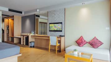 Una habitación de hotel moderna con suelo de madera, un sofá con cojines rojos, una mesita con una planta, un televisor montado en la pared y una zona de kitchenette.