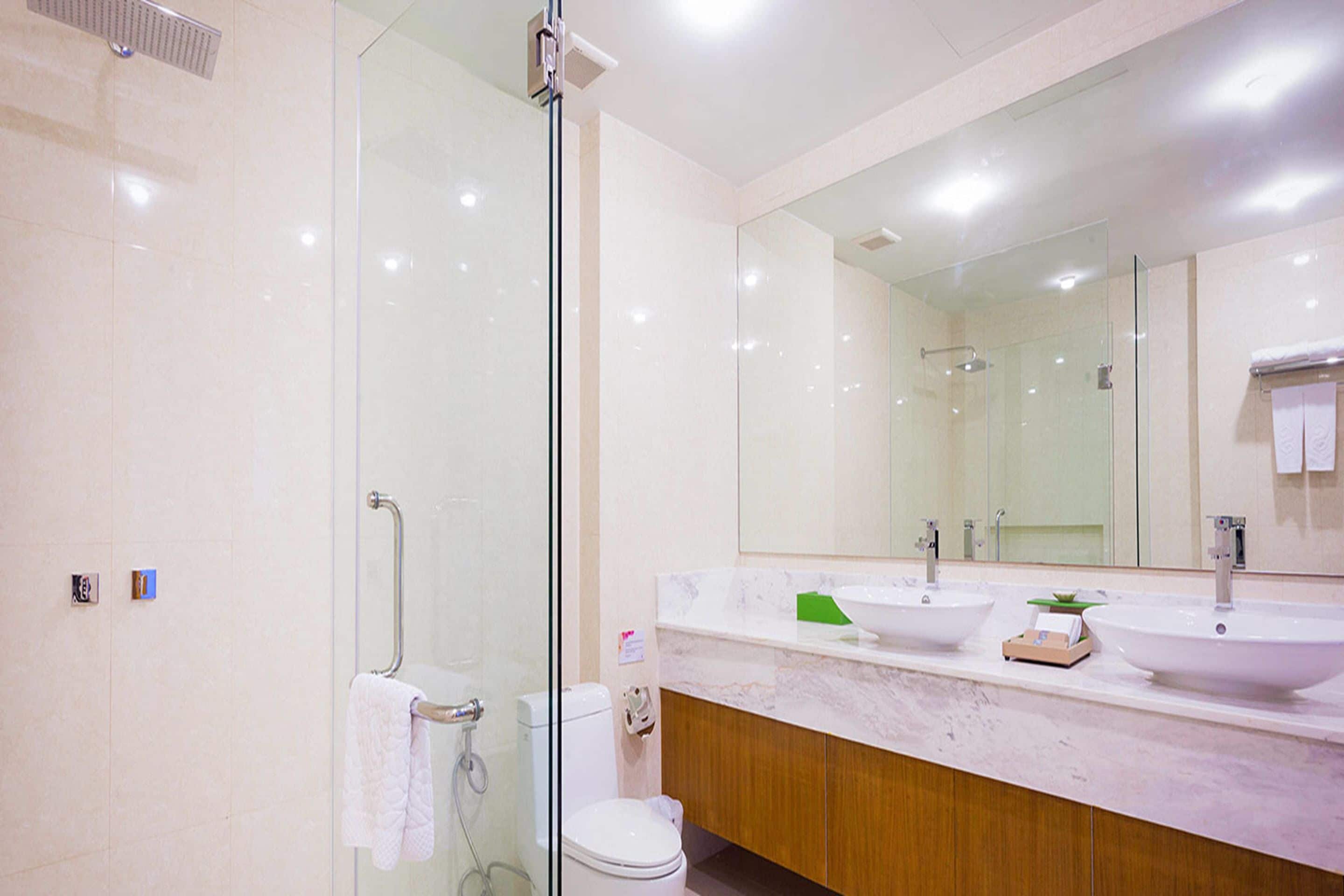 Un baño moderno con cabina de ducha de vidrio, inodoro y tocador con dos lavabos y un espejo grande.