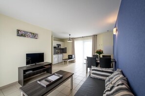 A modern living room with a flat-screen TV, a sofa, a dining table with chairs, and a kitchen area in the background. - Les Cottages du Saleys (Salies-de-Bearn)