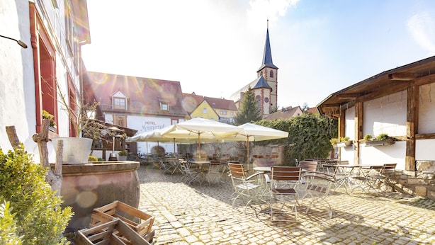 A cobblestone courtyard with outdoor seating, a church spire in the background, and a hotel sign.