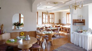A spacious dining area with multiple wooden tables and chairs, a chandelier, and a large window providing natural light.