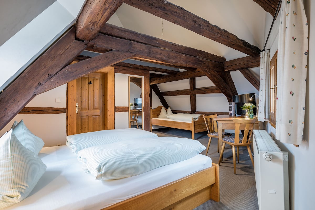 A bedroom with a wooden bed, a desk, and a chair. There is a window with curtains and a radiator on the wall.