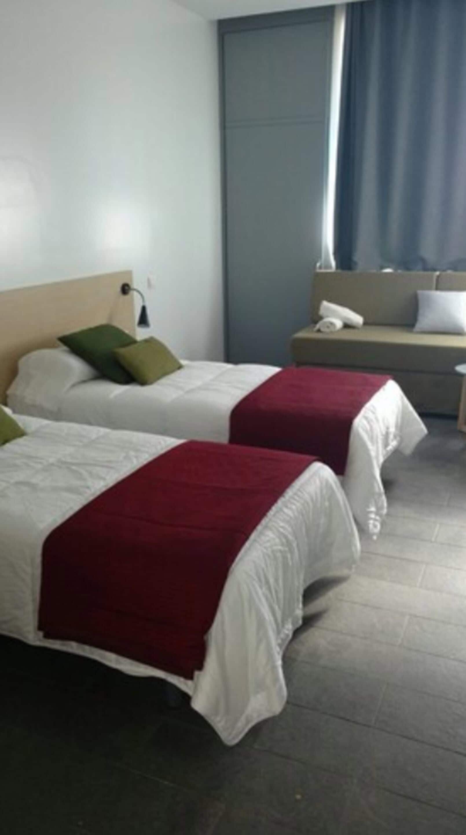 Habitación de hotel con dos camas, cada una con ropa blanca y una manta roja. Se ve un mesita de noche con una lámpara y un teléfono. La habitación tiene una cortina azul y una ventana con vistas al exterior.