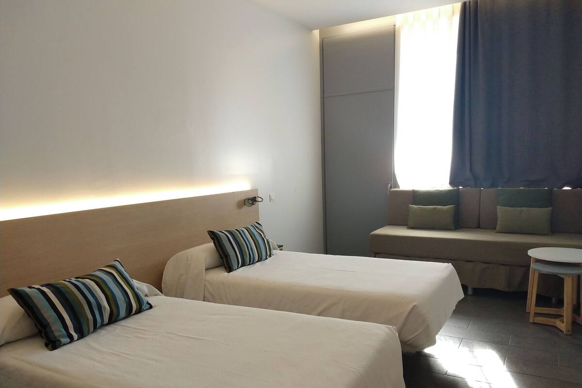 A hotel room with two beds, a sofa, a small table, and a window with curtains.