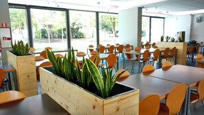 A spacious indoor dining area with multiple tables and chairs, large windows offering a view of greenery outside, and wooden planters with green plants. - Centre Esplai Hostel (El Prat de Llobregat)