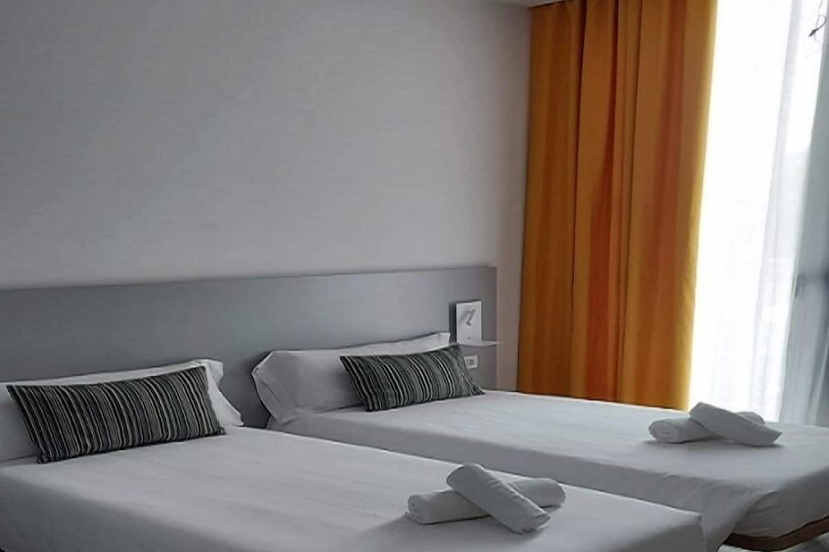 A hotel room with two single beds, white bedding, striped pillows, and a large window with orange curtains.