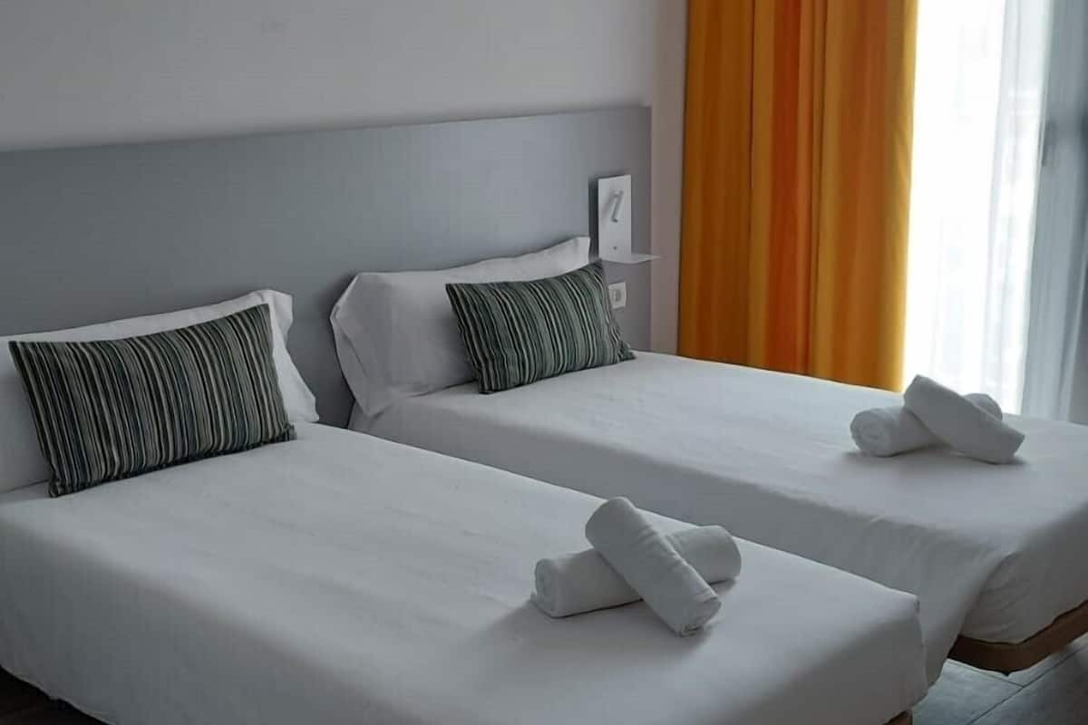A hotel room with two single beds, a wooden floor, and a large window with orange curtains.