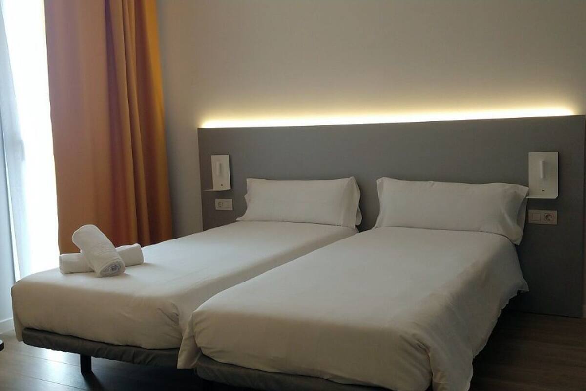 A hotel room with two beds, a bedside table, a mirror, and a window with curtains.