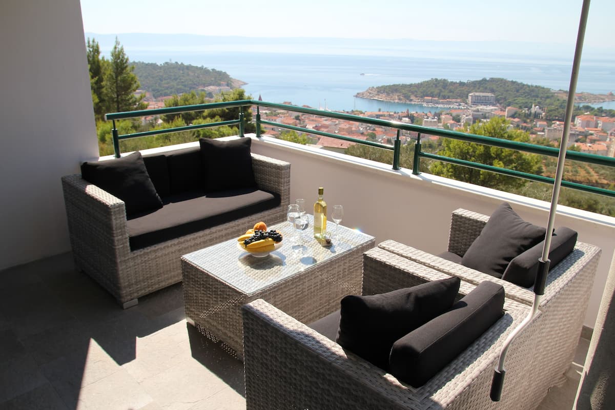 A balcony with wicker furniture, a table with a bottle and fruit, and a view of the sea and town.