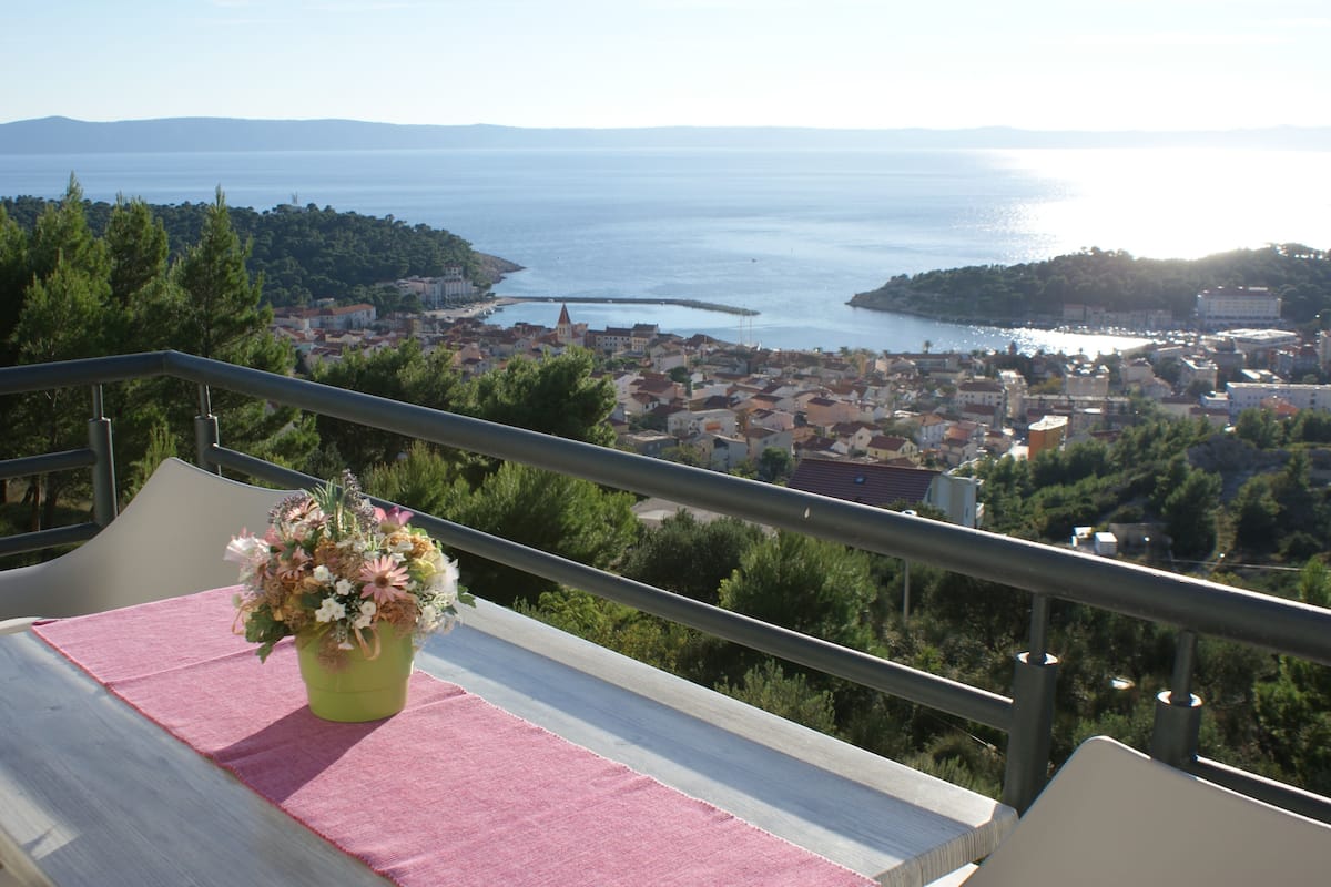 A balcony with a pink tablecloth and a vase of flowers, overlooking a coastal town with buildings and a body of water.