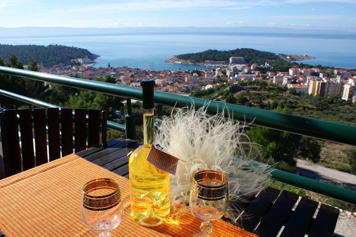 A table with a bottle of wine, two glasses, and a decorative feather arrangement on a balcony overlooking a coastal town.