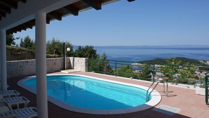 Piscina com vista para o mar, rodeada por um terraço coberto com cadeiras brancas.