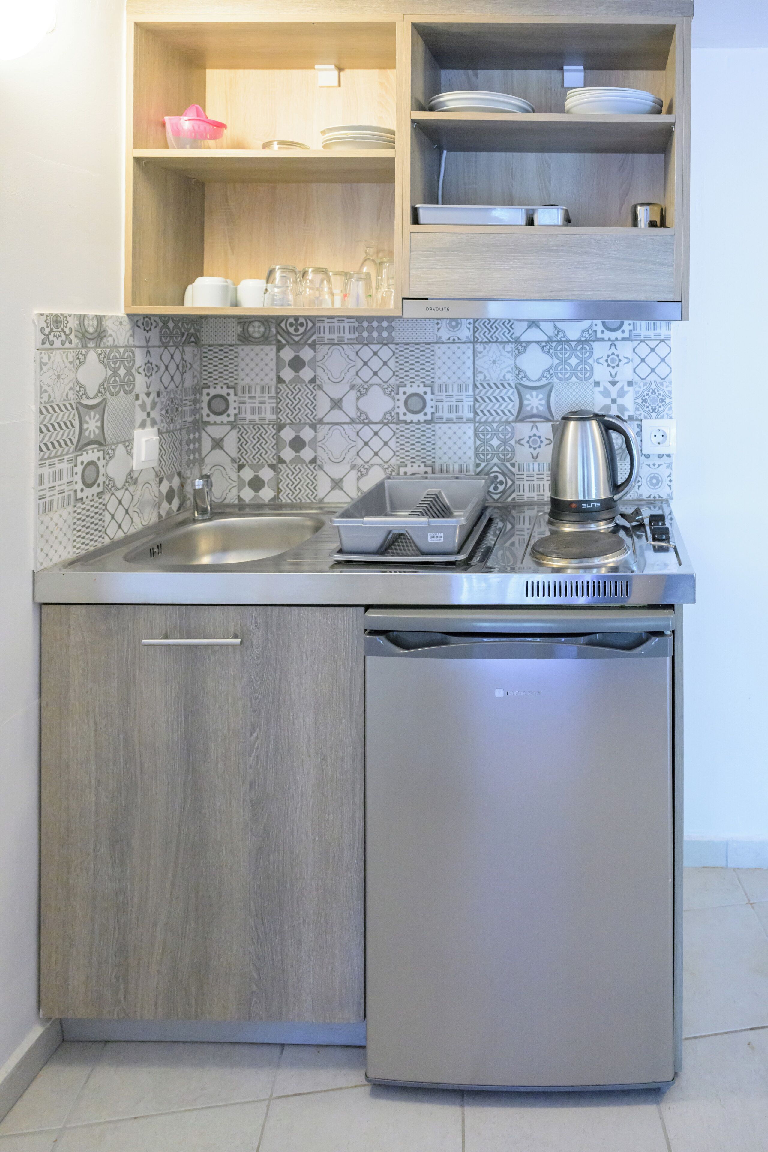 A compact kitchen with a built-in dishwasher, a kettle, and a variety of glassware and dishes on the shelves.