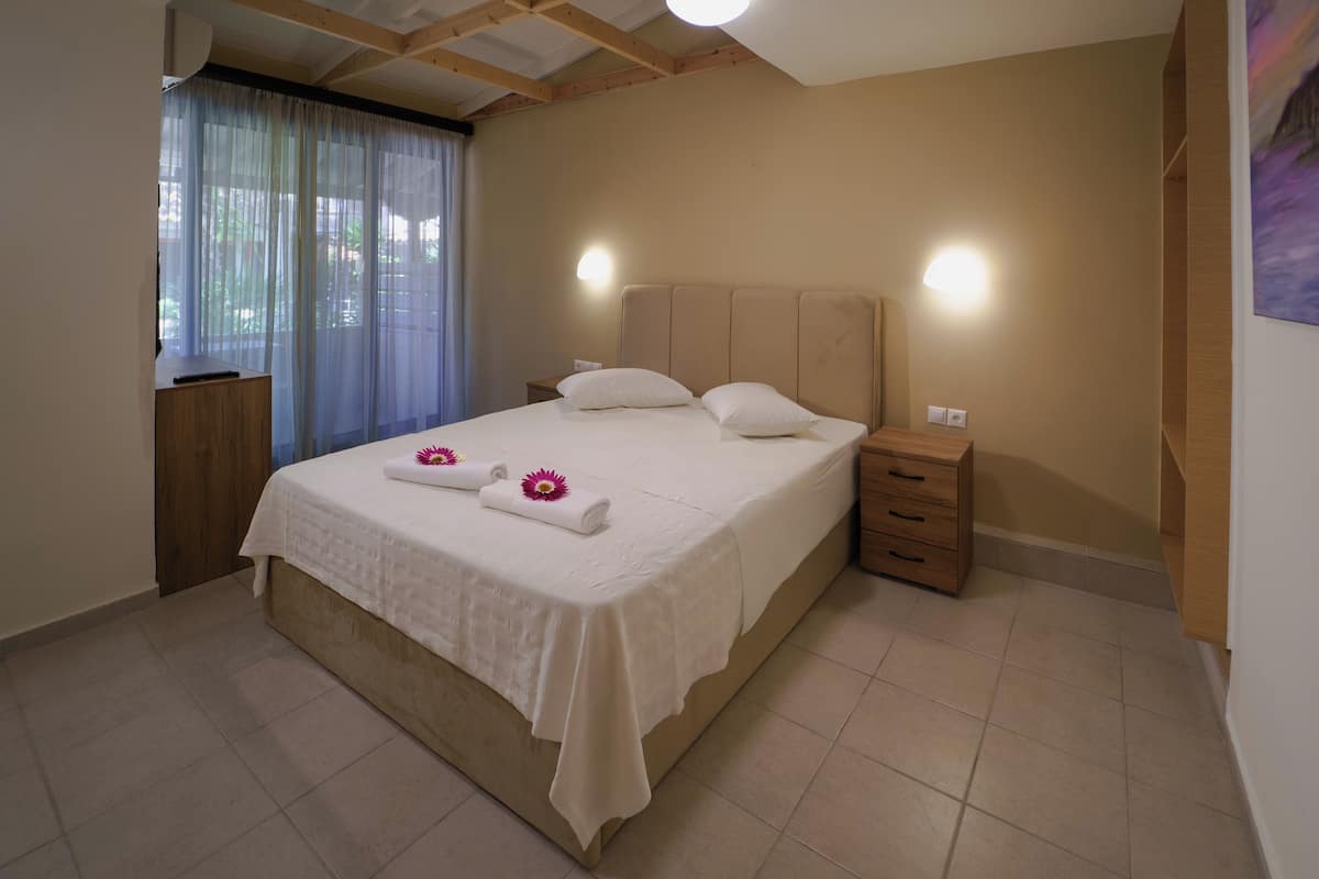 A neatly made bed with white linens and two pillows, a bedside table with a lamp, and a window with sheer curtains.