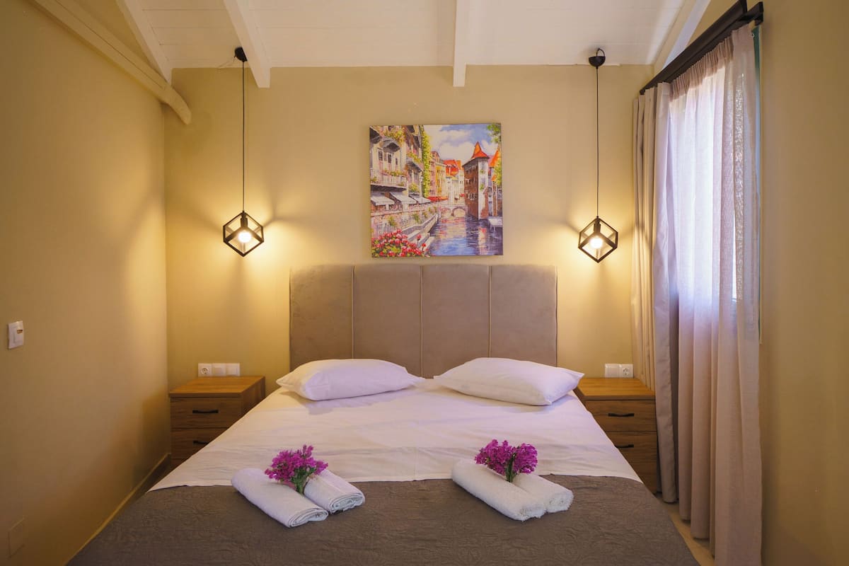A neatly made bed with a brown bedspread, two white pillows, and a decorative towel with flowers. There are two bedside lamps, a painting on the wall, and a window with curtains.