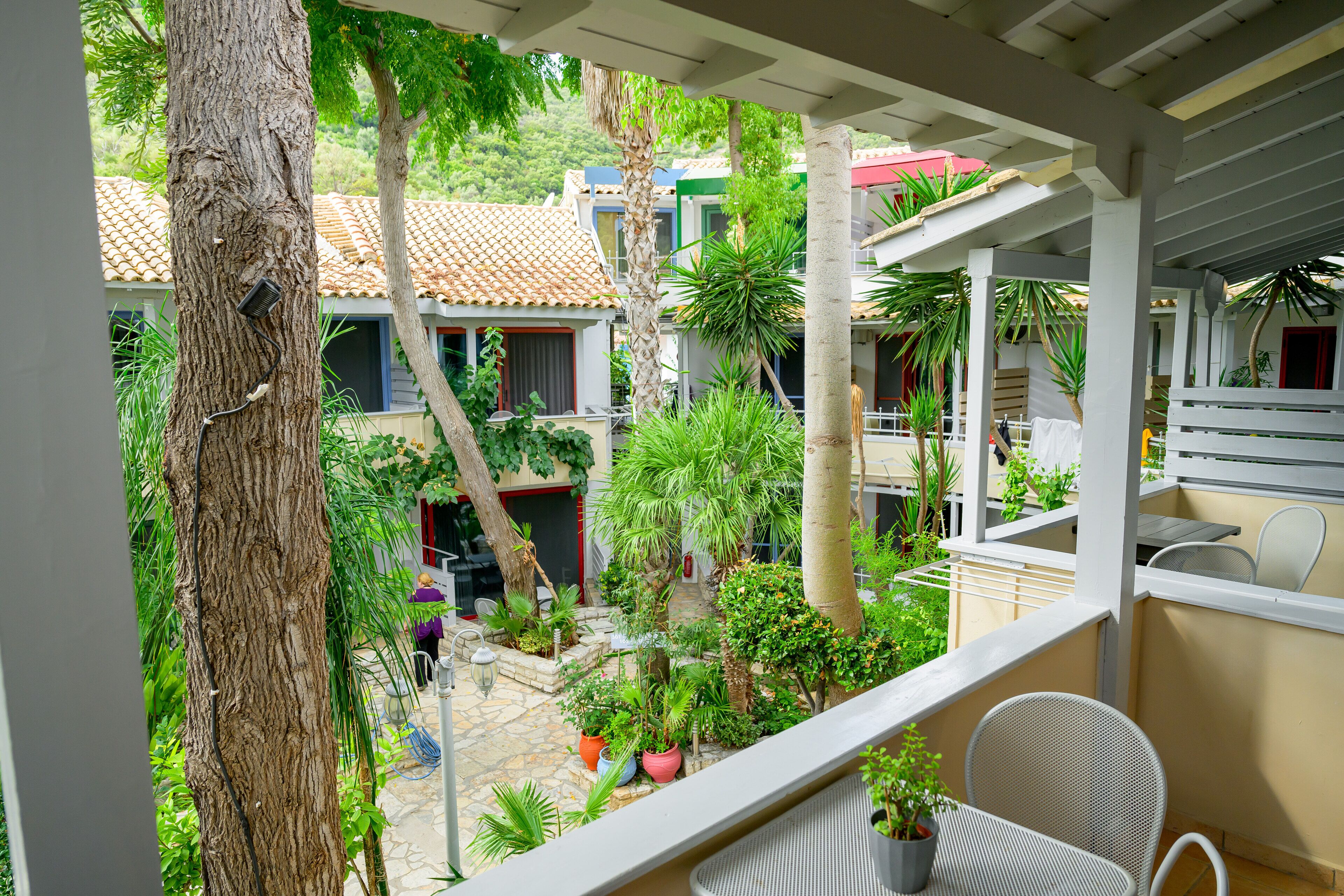 A balcony with wicker furniture, potted plants, and a view of a courtyard with trees and a building with a tiled roof.