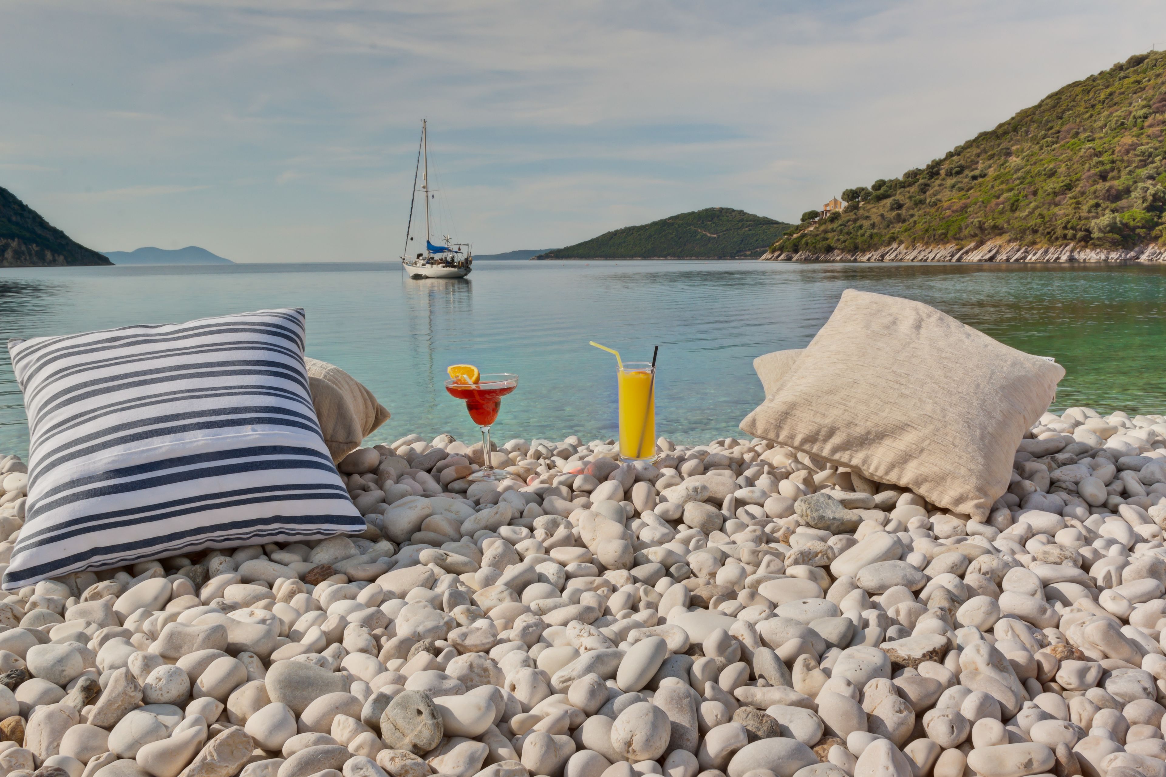 A pebbled beach with a sailboat in the distance, pillows, and a drink on the shore.