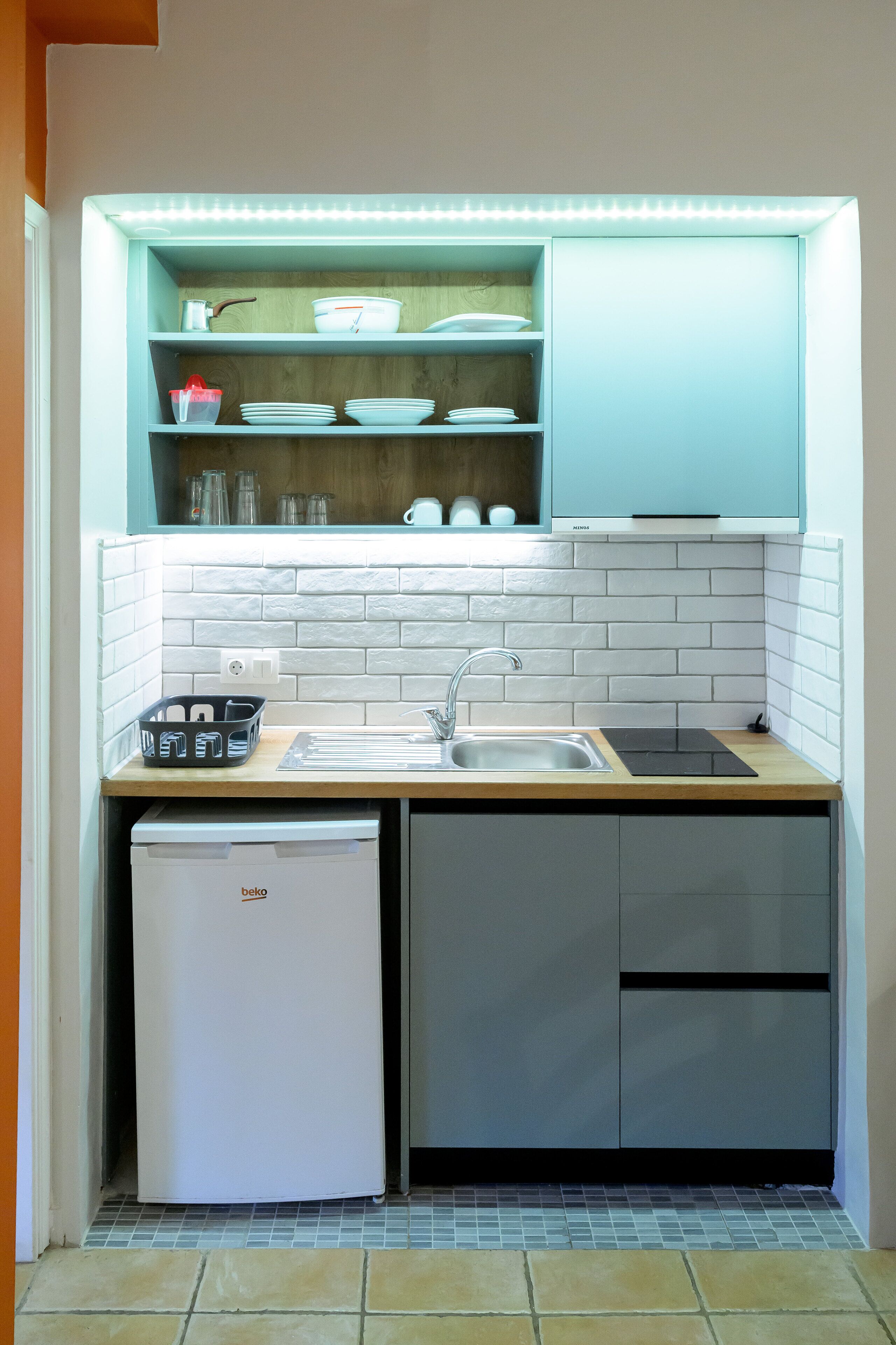 Una cocina compacta con pileta integrada, un refrigerador pequeño y una alacena sobre la encimera con vajilla y vasos.