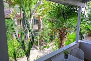 A balcony with a view of a garden, including cacti and palm trees, and a table with a potted plant.