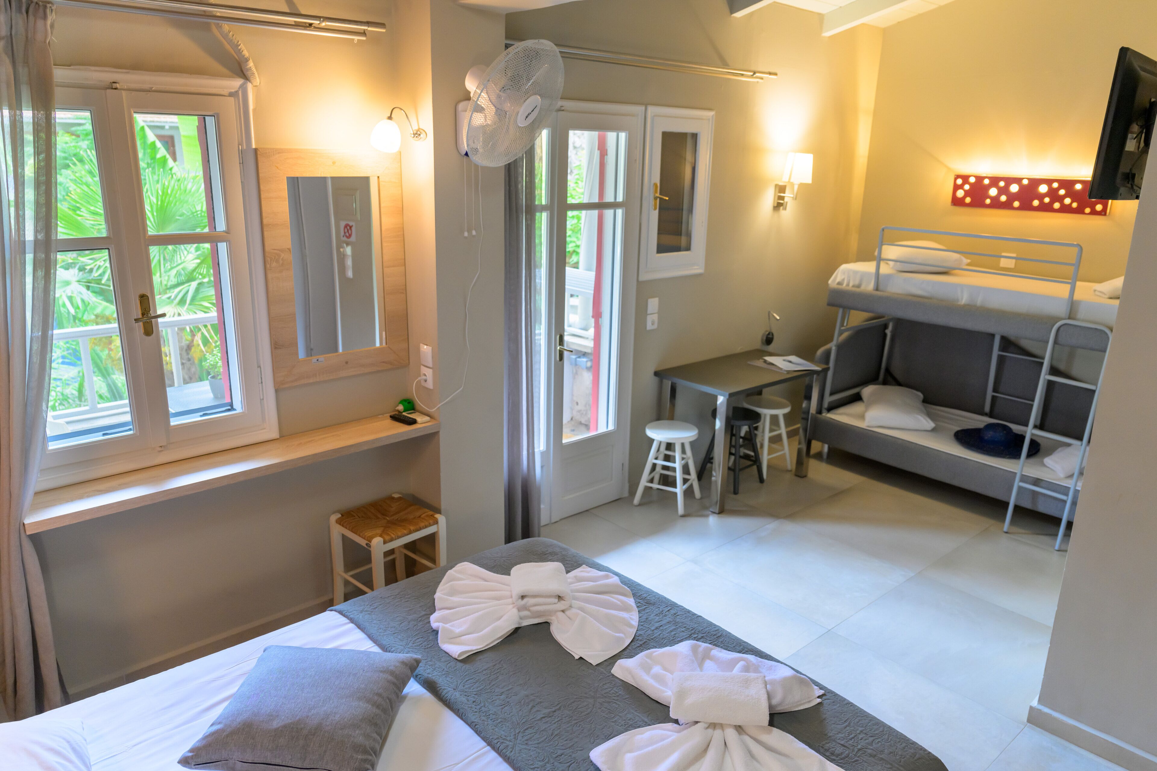 A hotel room with a bed, a desk, a bunk bed, a fan, and a view of greenery outside.