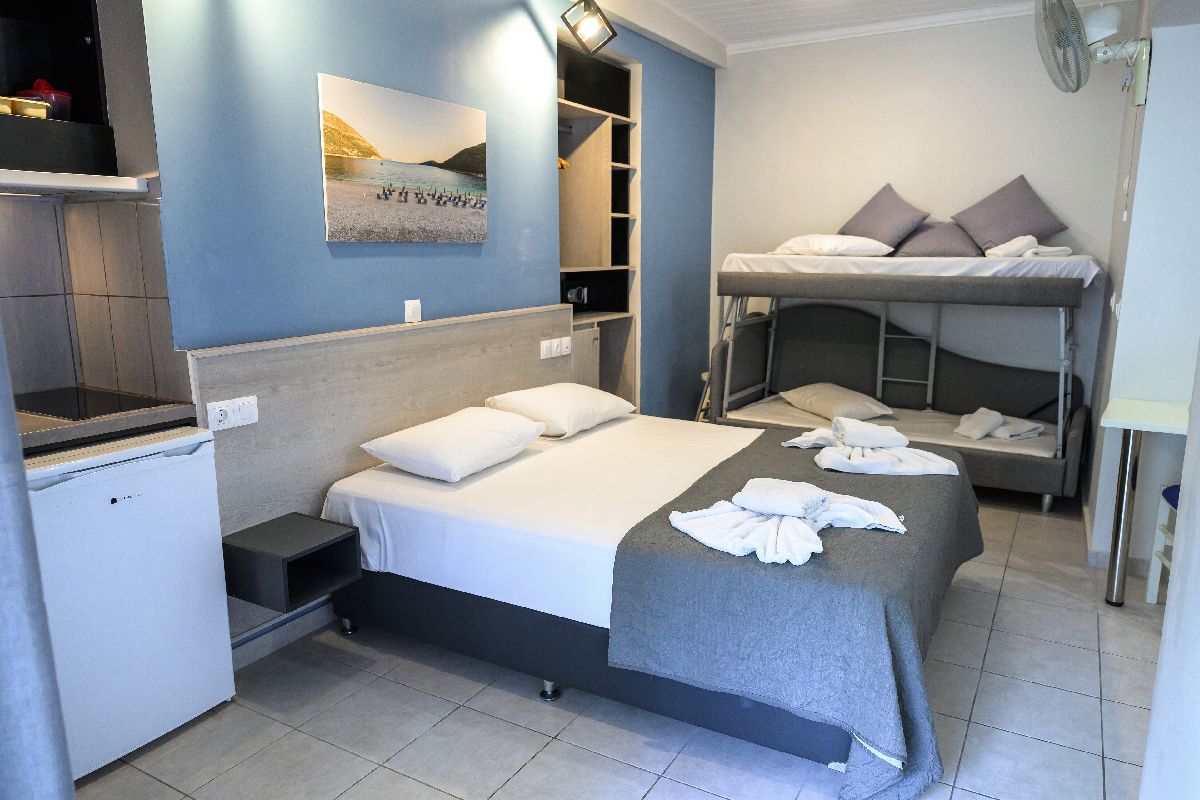 Une chambre d’hôtel avec un lit simple, un lit superposé, un petit réfrigérateur, une étagère avec quelques objets et un tableau au mur.