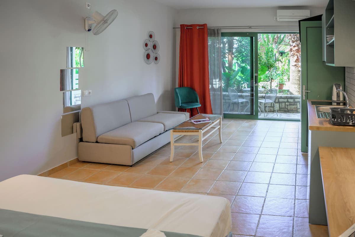 A modern hotel room with a bed, a sofa, a small table, a chair, a kitchenette, and a sliding glass door to an outdoor area with plants.