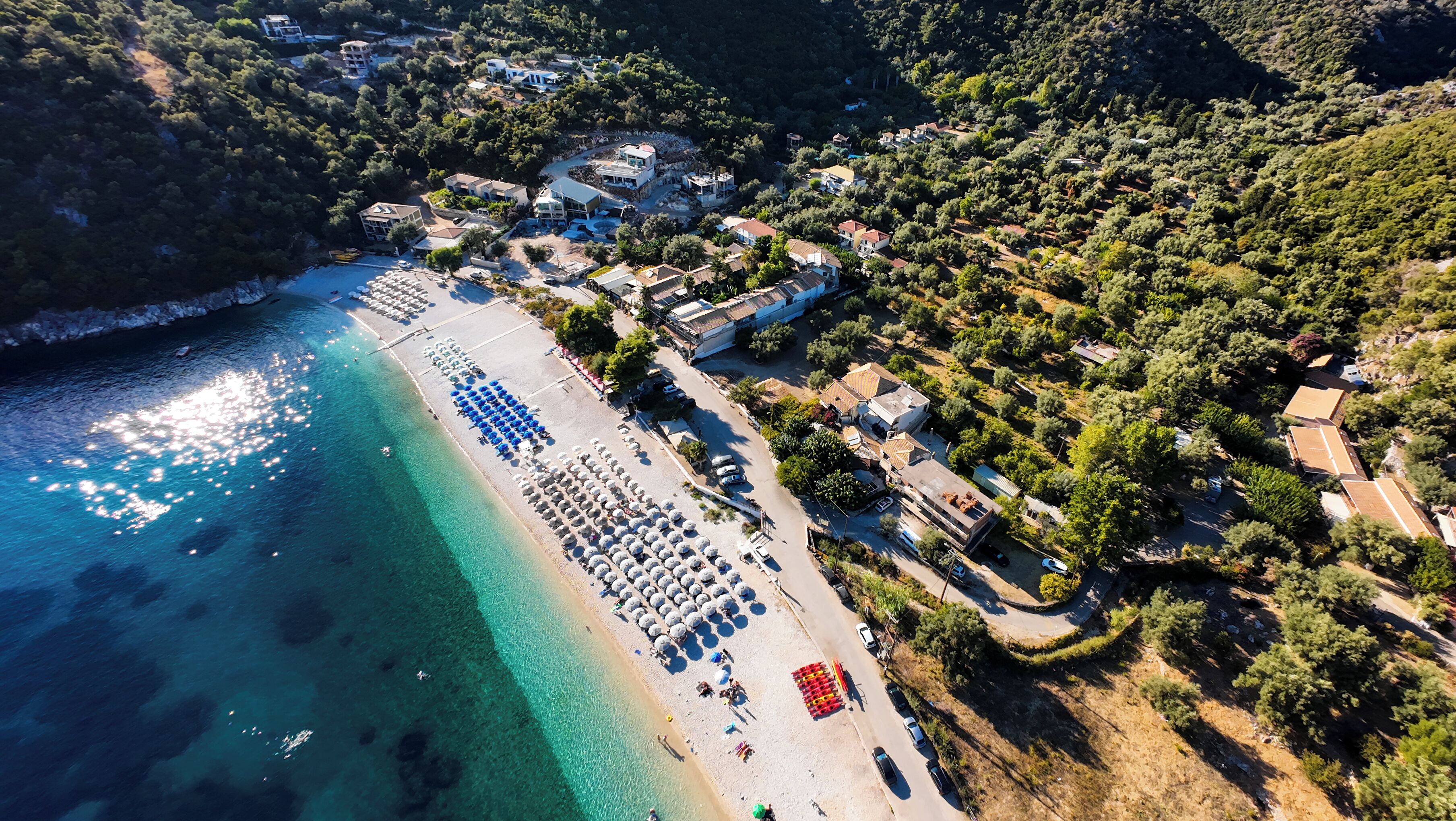 An aerial view of a coastal area with a beach, clear turquoise waters, and a row of houses with a hilly backdrop.